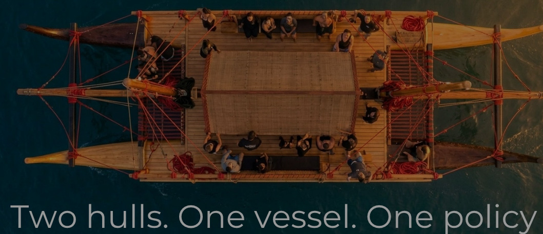 A double-hulled waka hourua under way, seen from directly above — two hulls, a shared deck, and crew at work